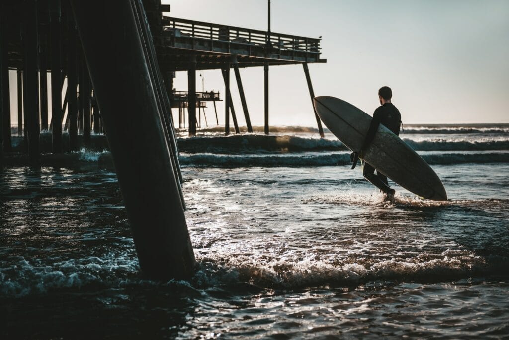 A surfer carrying a surfboard near a Pismo Beach pier.