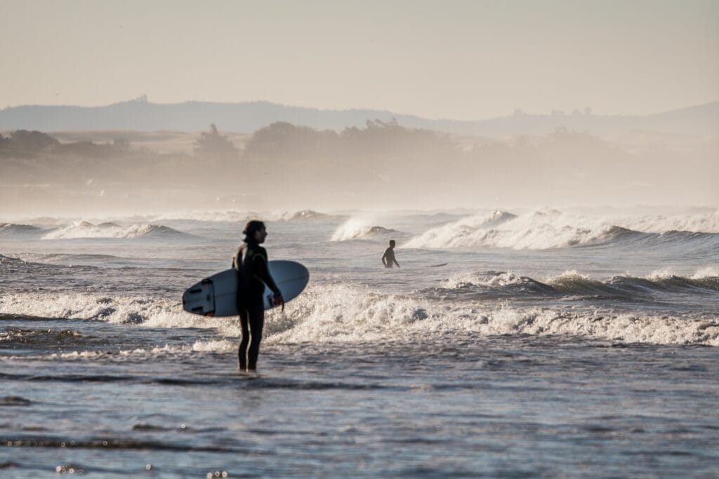 A man in a wetsuit standing in the ocean at Pismo Beach with a surfboard.